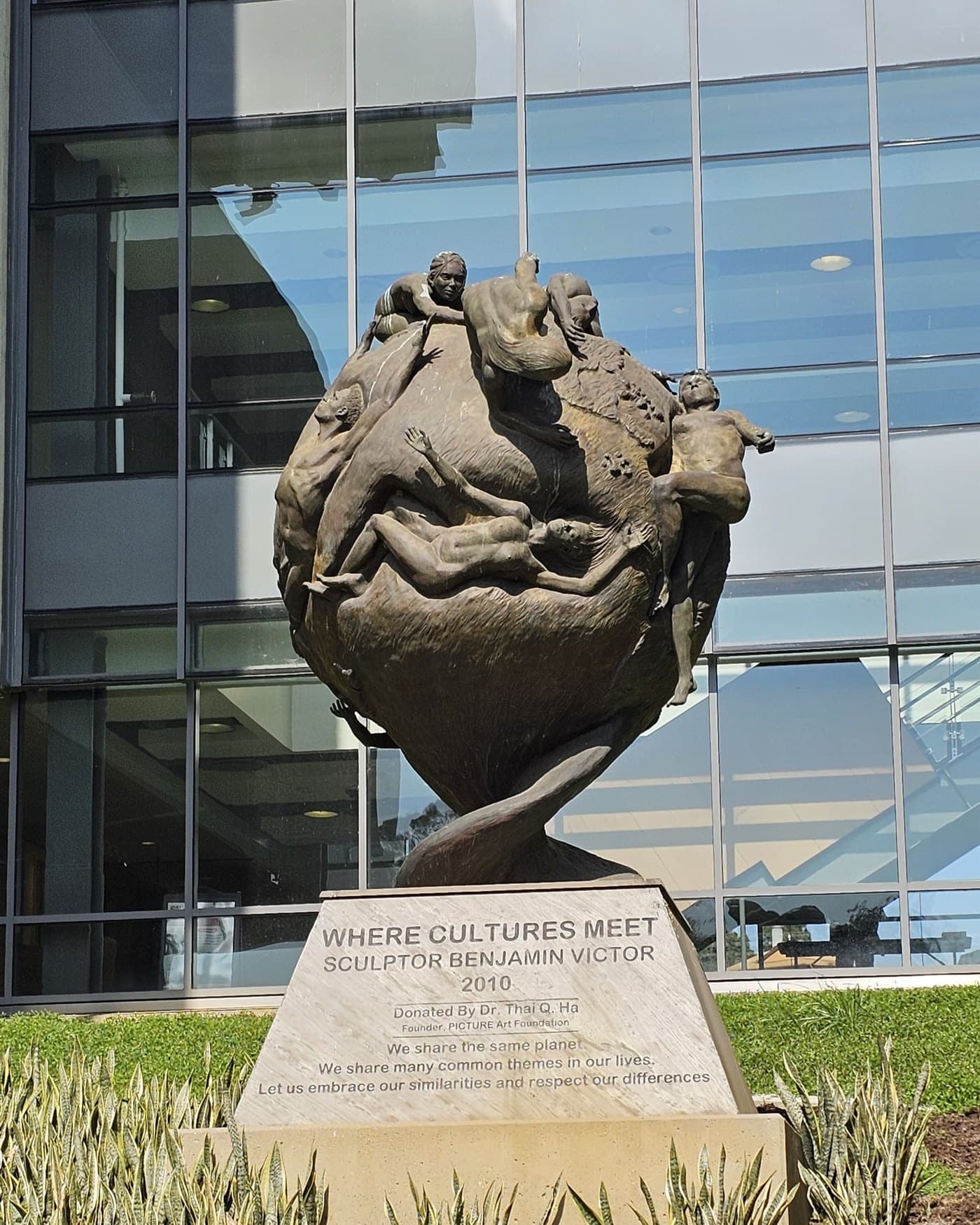 Where Cultures Meet: a bronze sphere of figures reaching across a globe, mounted on a stone plinth outside a glass-fronted university building