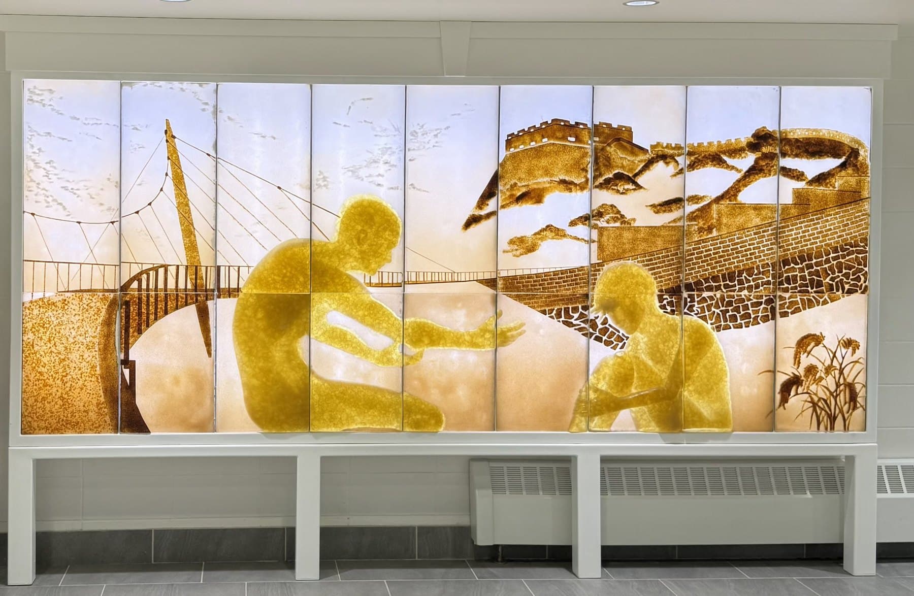 Further the Tradition: two amber glass figures reaching toward one another across panels of pale glass, a suspension bridge behind one and the Great Wall of China behind the other